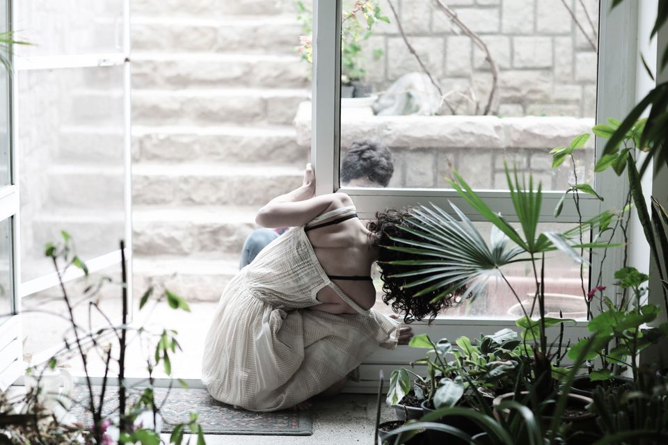 A woman in a white dress folds herself against a glass door among plants while a man waits on the other side