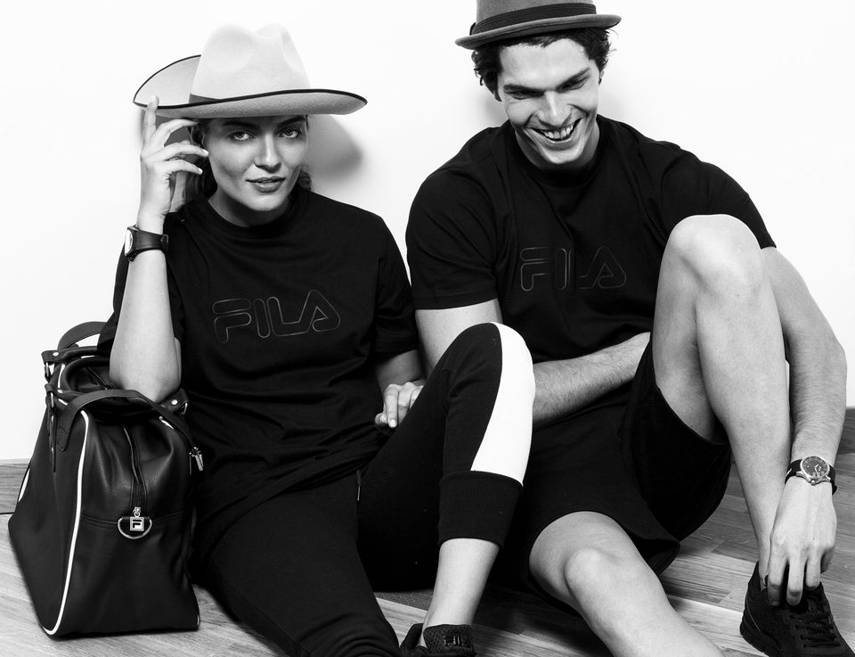 Two models in black FILA outfits and hats sit on the floor laughing, black and white