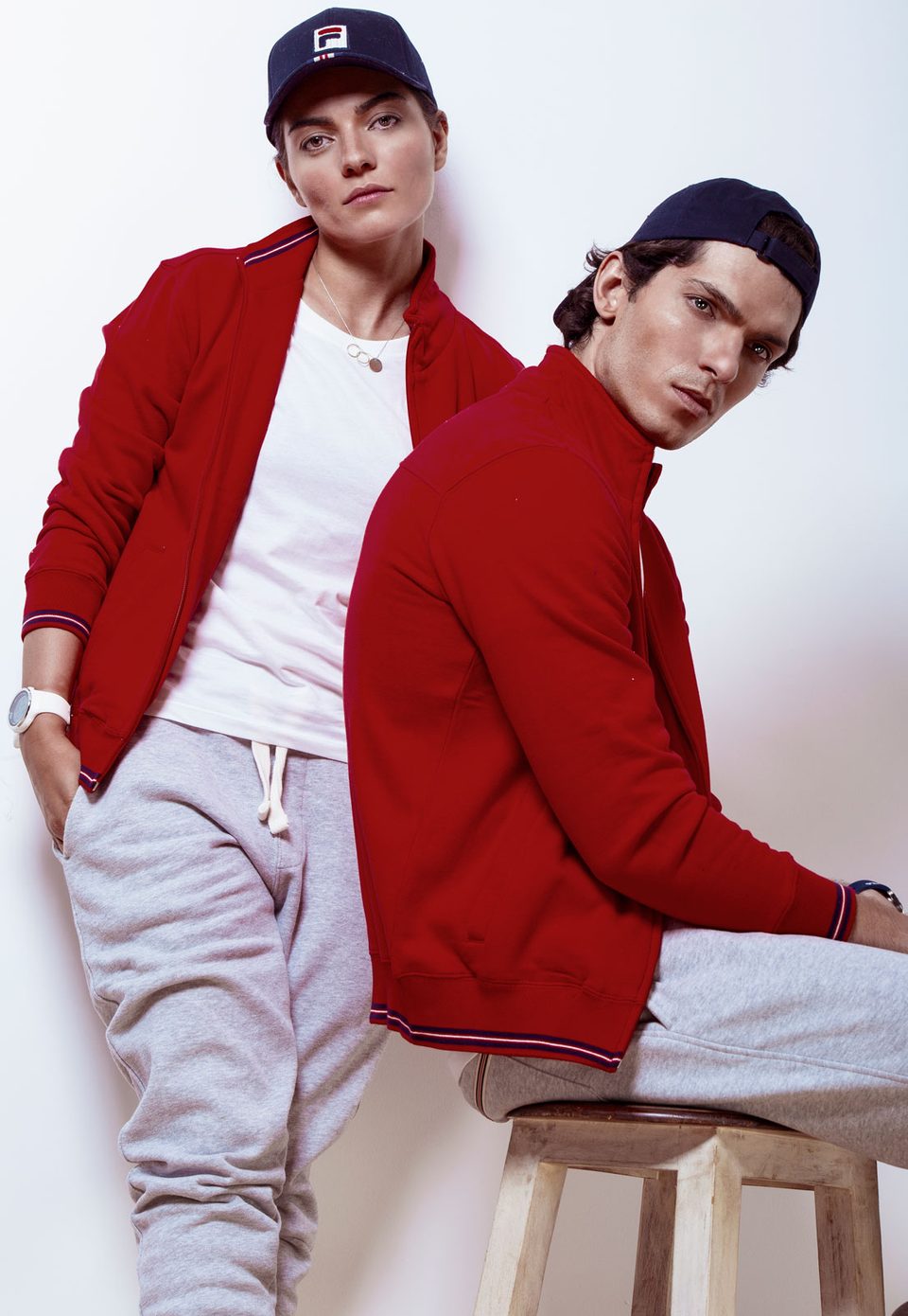 Two models in red FILA track jackets and navy caps pose against a white wall