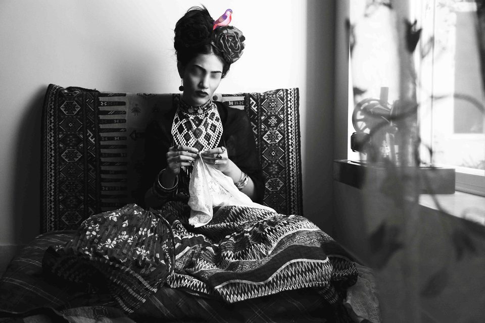 The Fridah figure embroiders on a kilim-covered sofa beside a bright window, black and white