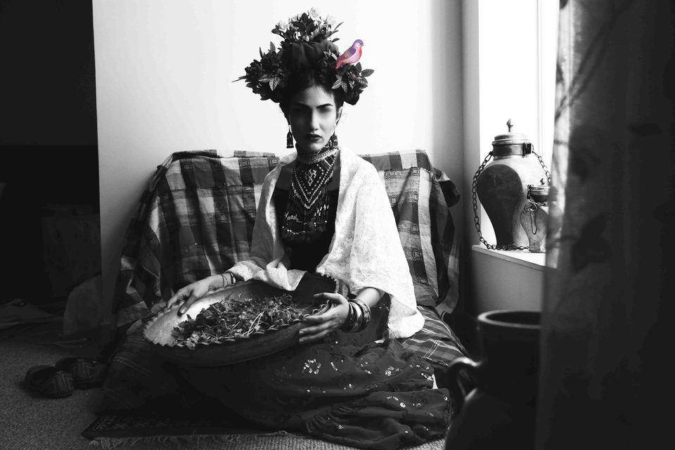 A woman styled as Frida Kahlo, with a bird in her braided hair, sits on a patterned kilim sofa in black and white