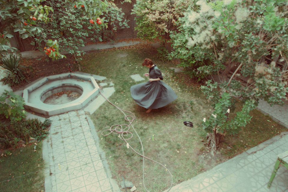 Seen from above, a woman in a long skirt turns in a courtyard garden beside an octagonal pool