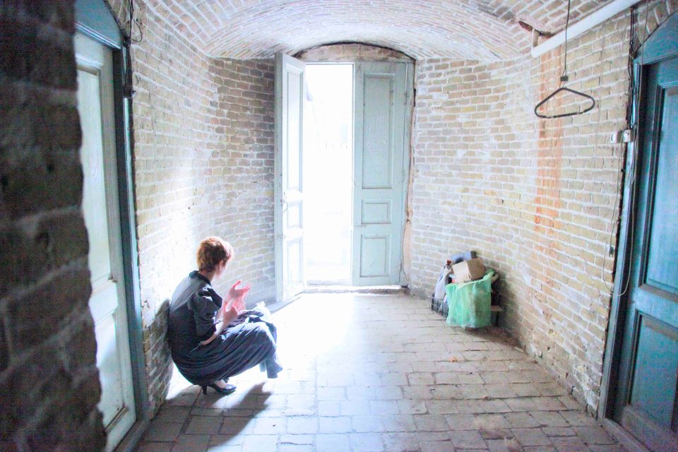 A figure crouches in the light of an open doorway inside a brick-vaulted room in an old house in Mashhad