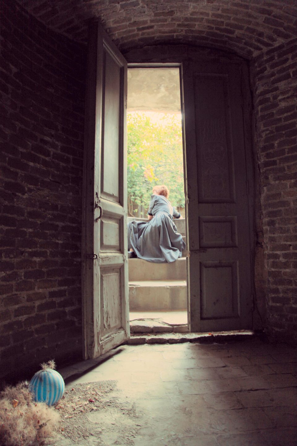 A seated figure framed by tall wooden doors that open onto a sunlit garden
