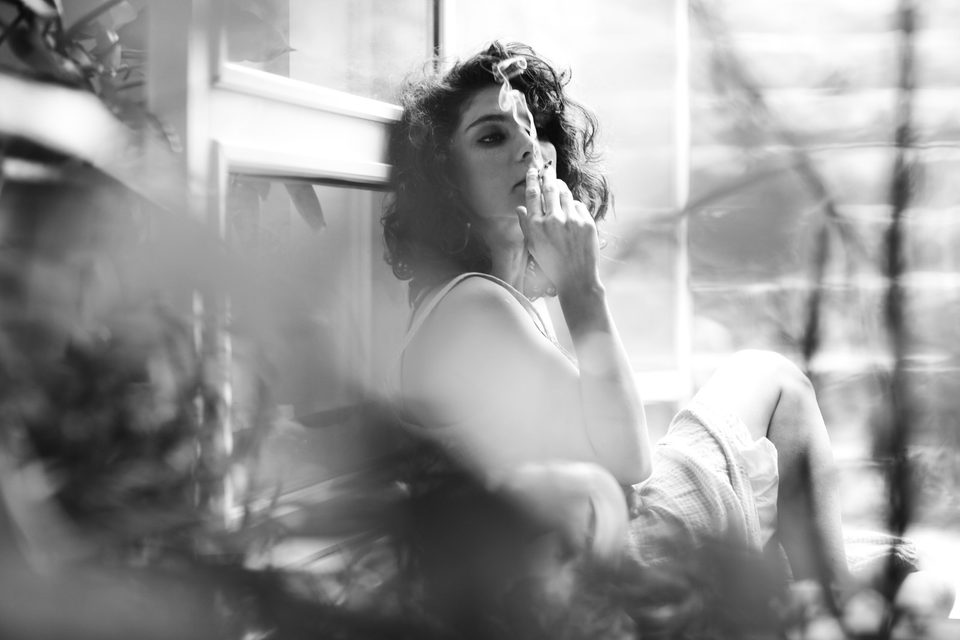 A woman reclines by a window, plants and haze softening the foreground, black and white