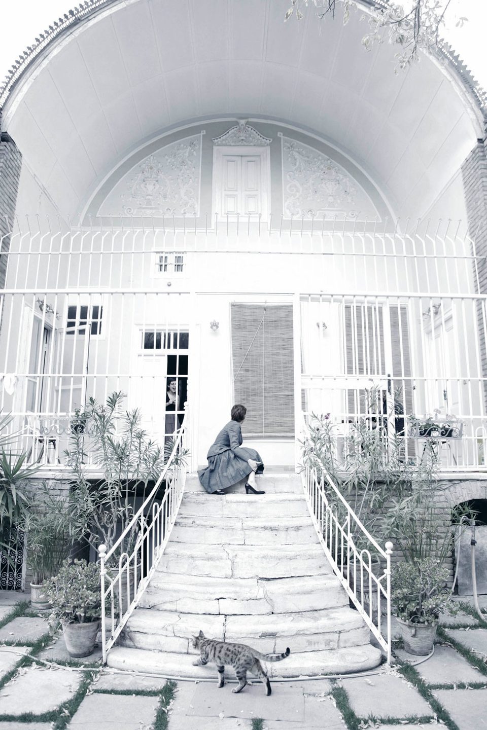 A figure sits on the white steps of an old house with arched windows while a cat walks below