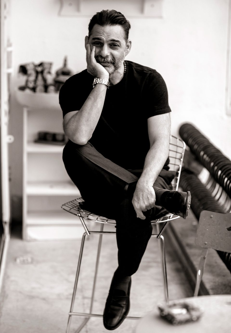 Payman Maadi seated on a wire chair in a studio, hand against his cheek, black and white