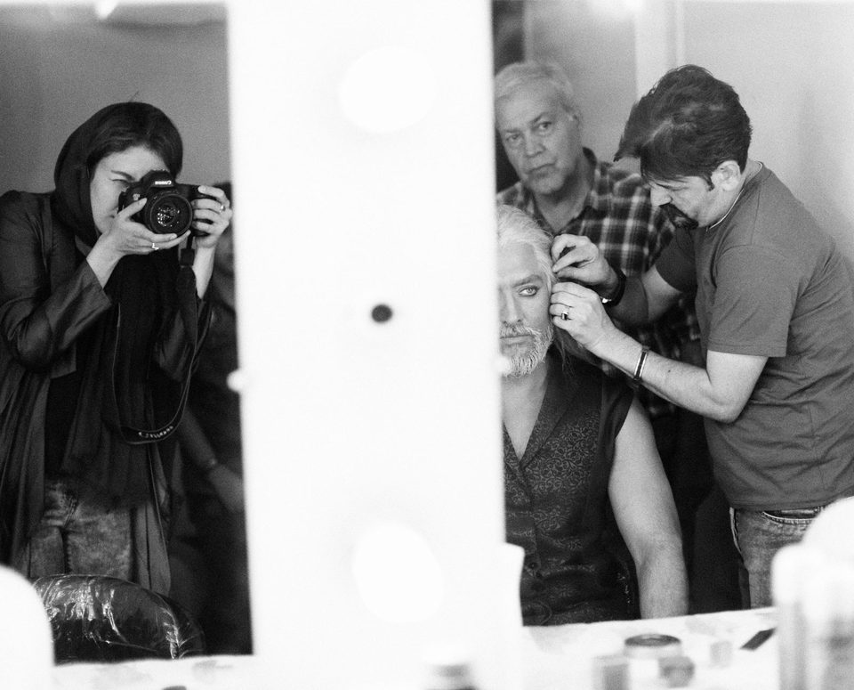 A mirror reflects a woman holding a camera beside the makeup team at work backstage