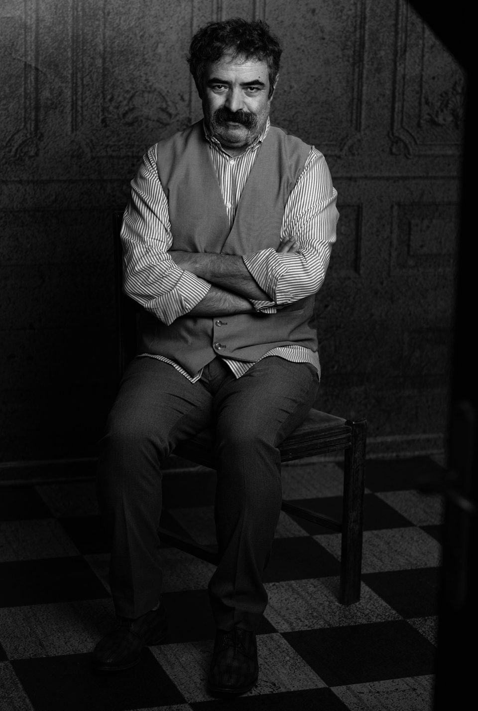 Actor Hassan Majooni seated in a stone hall with his arms folded, black and white