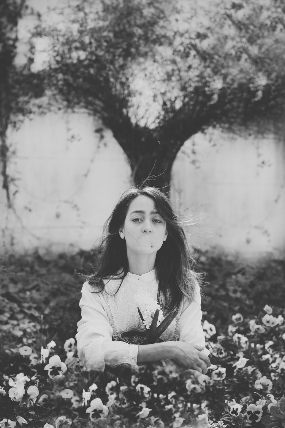 A young woman lies among flowers beneath a tree and looks up at the camera, black and white