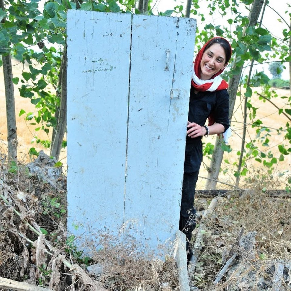 Hedieh Tehrani in a red headscarf smiling from behind a pale blue wooden door, trees behind her