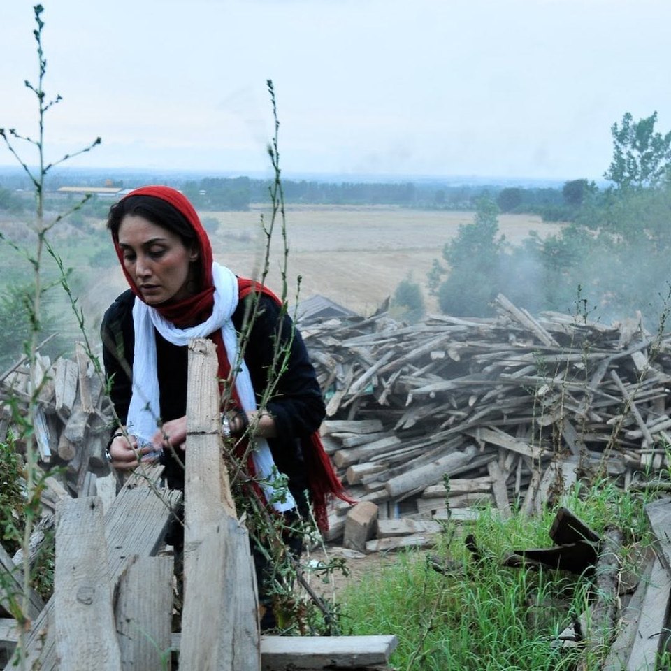 Hedieh Tehrani in a red headscarf and white scarf beside a pile of old timber, fields and a misty horizon behind her