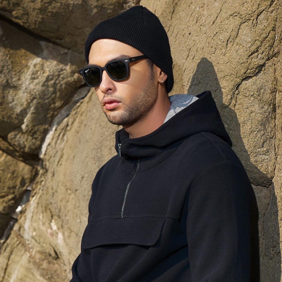 A man in a black hoodie, beanie and sunglasses leans on a rock face