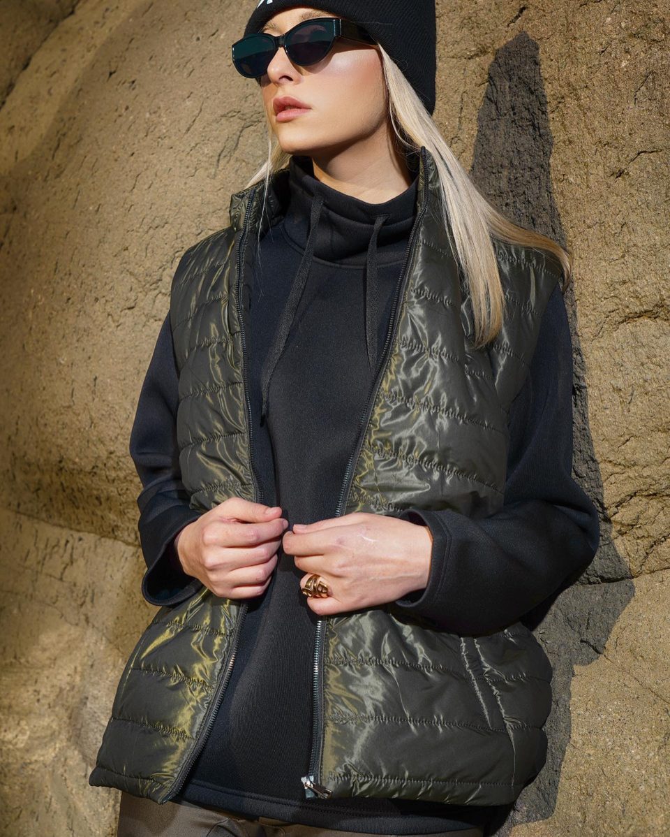A woman in sunglasses and a black beanie wears an olive quilted vest against rock