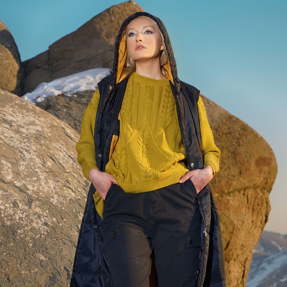 A model in a mustard cable-knit sweater and black vest stands against sandstone rocks, Adora campaign by Reihanet Studio