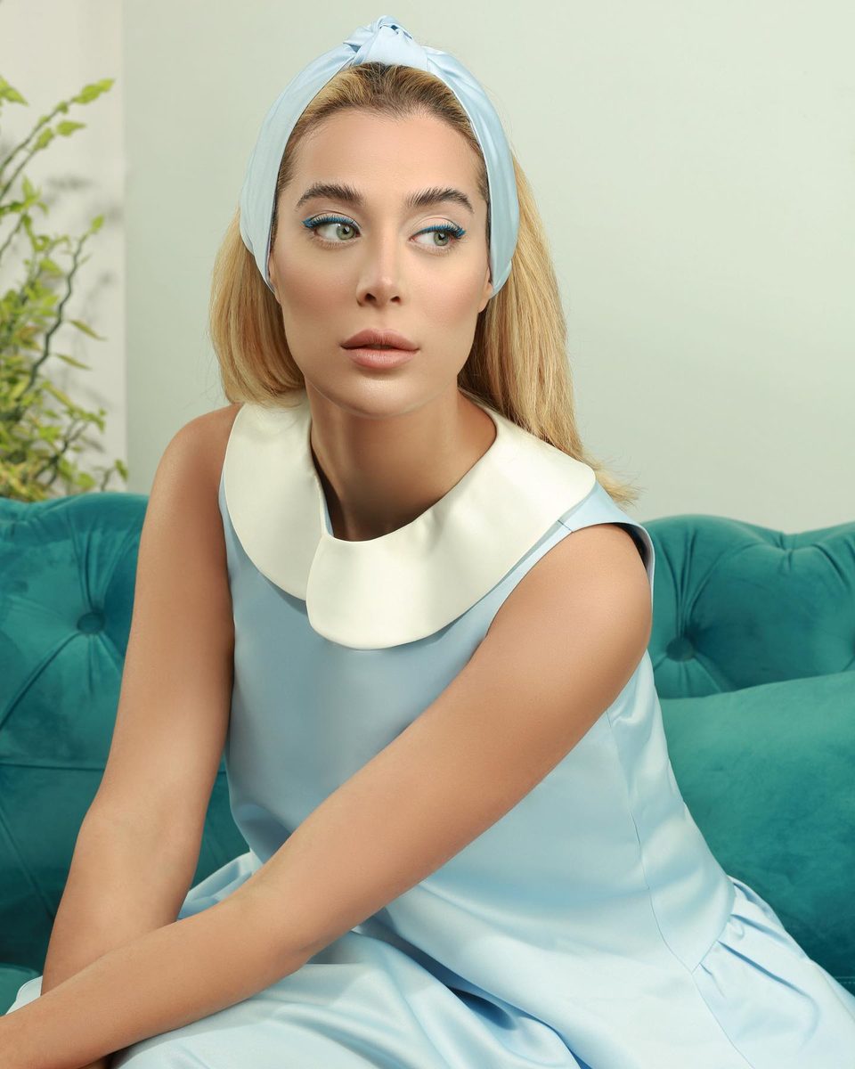 A woman in a pale blue dress with a white collar and blue headband sits on a green sofa