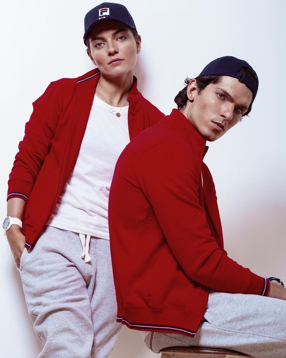 Two models in red FILA track jackets and navy caps pose against a white wall
