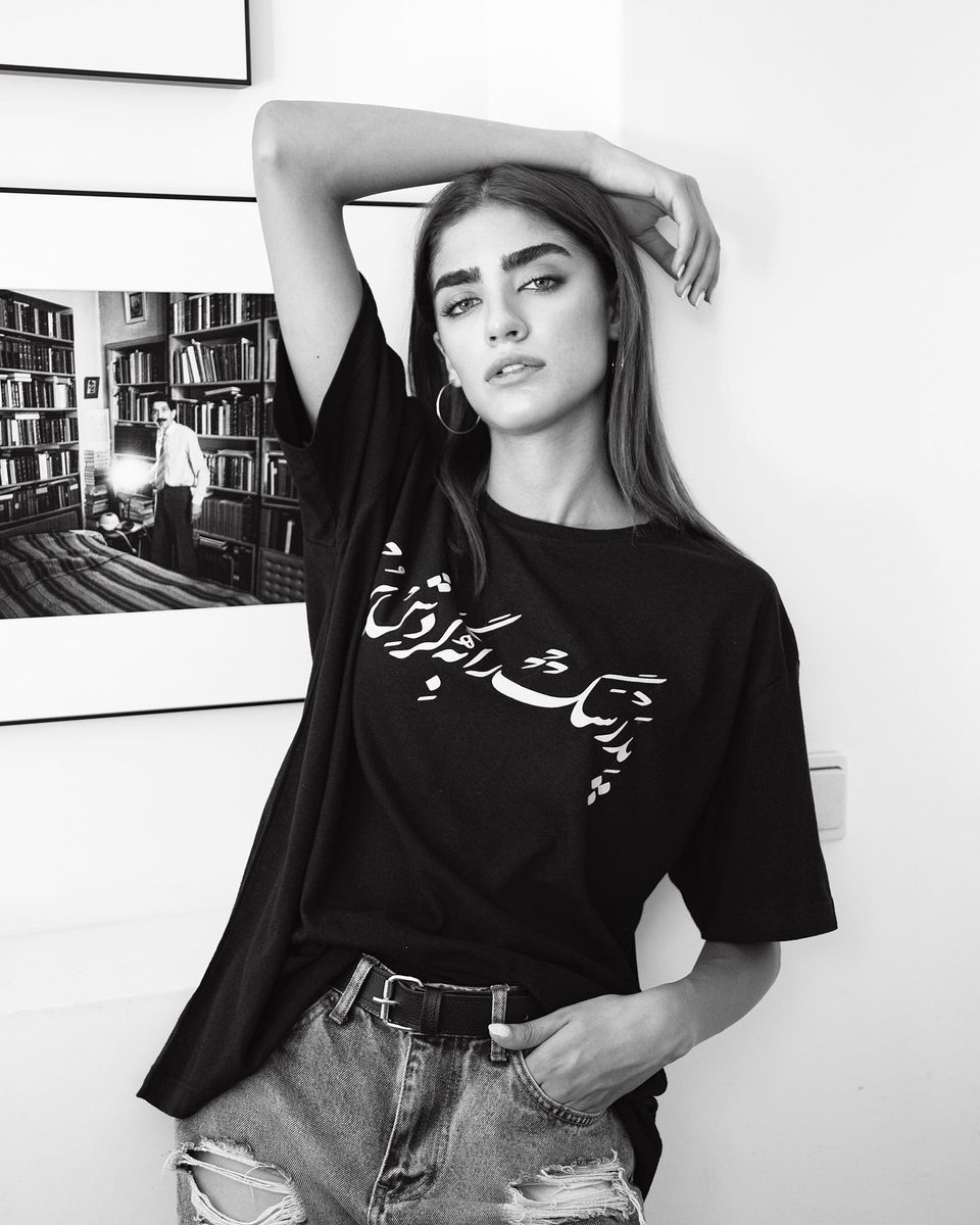 Black-and-white photograph of a model in a T-shirt with Persian script, hand in her hair
