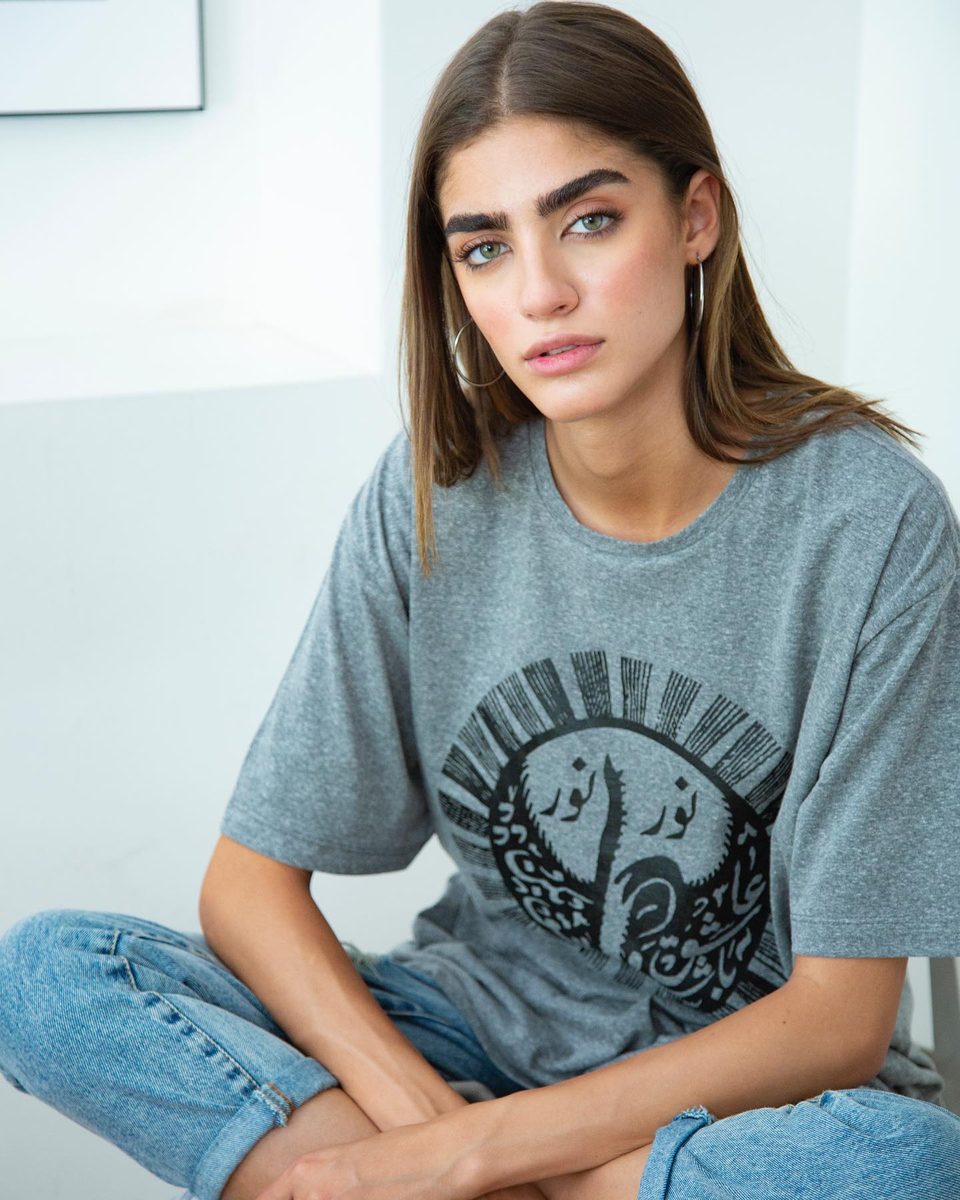 A model sits cross-legged in a grey T-shirt printed with a Persian calligraphy design