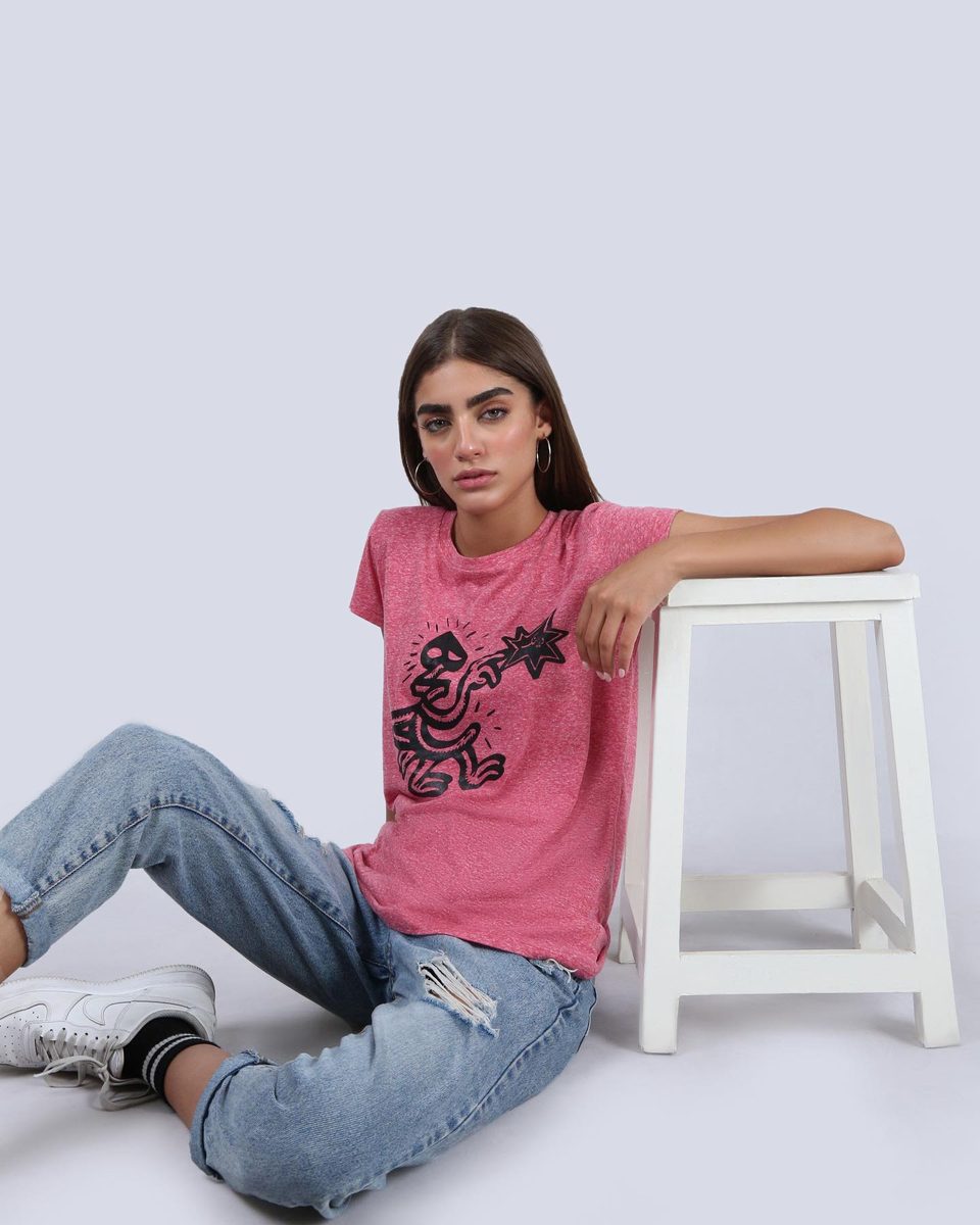 A model in a pink printed T-shirt and ripped jeans leans on a white stool