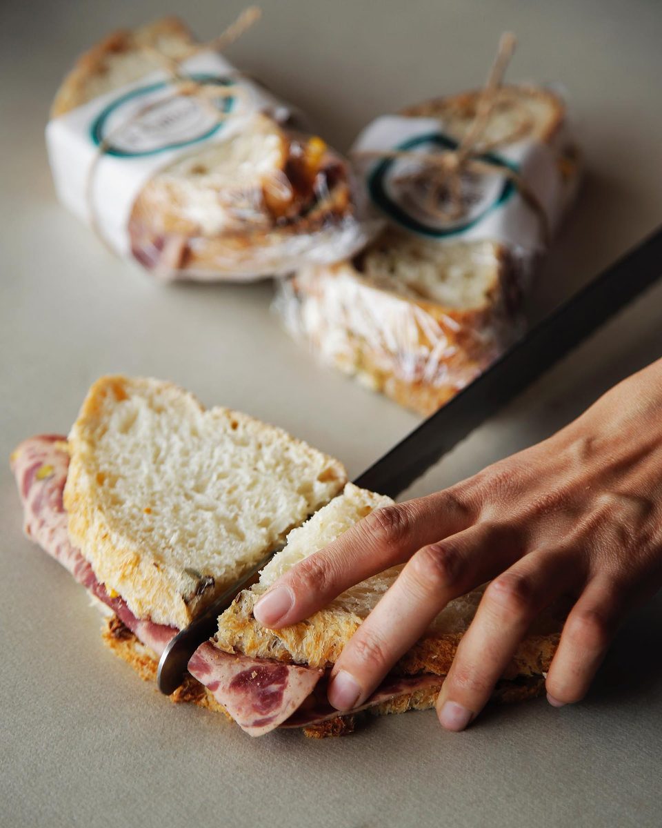 Hands slice a filled sandwich next to wrapped pastries