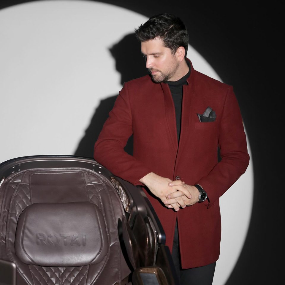 A man in a burgundy blazer leans on a Rotai massage chair under a spotlight