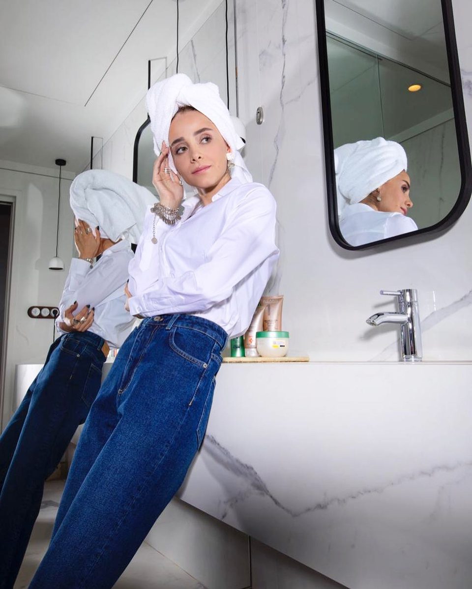 A woman with a towel on her head leans on a marble washbasin in white shirt and jeans
