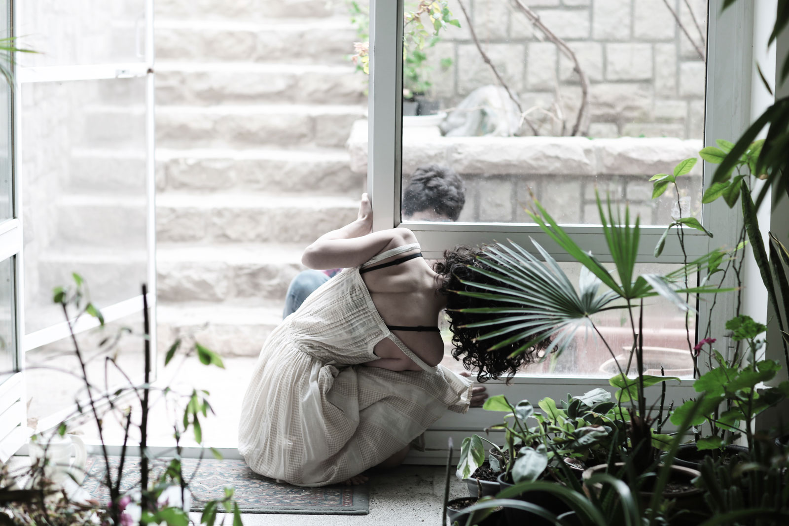 Whisper through the Glass – Mirrors and Women, 2013 A woman crouching beside a glass door surrounded by houseplants, photographed in soft natural light - part of the “Mirrors and Women” collection, 2013.